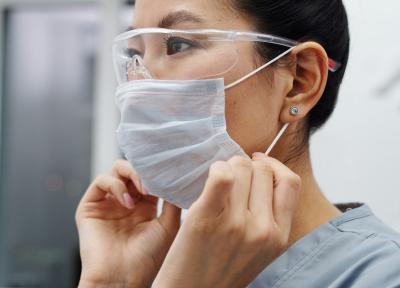 Woman in scrubs and eye shield adjusting straps on a face mask.